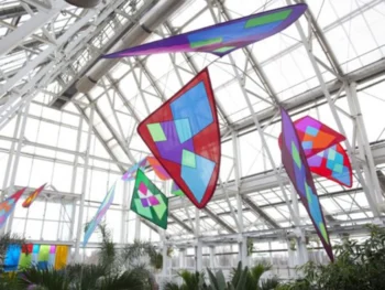 Colorful geometric kites hang from the ceiling inside a bright greenhouse with large windows and plants below.