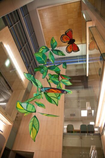 Large butterfly and leaf sculptures hang from the ceiling in a modern, multi-story atrium with glass and wood panels.