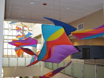 Colorful fabric sculptures hang from the ceiling in an indoor atrium with glass railings and large windows.