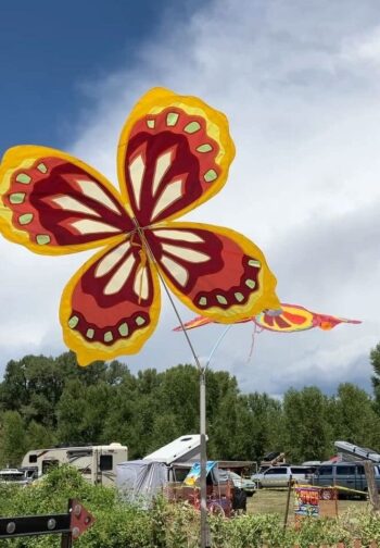 Large, colorful butterfly-shaped kite flying outdoors above tents and vehicles under a partly cloudy sky.