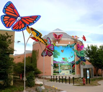 Colorful butterfly sculptures and a mural decorate the exterior of a circular building on a sunny day.