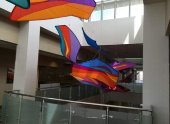 Colorful abstract kites hang from the ceiling in a bright, modern building with glass railings.