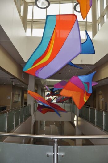 Colorful abstract kite-like sculptures hang from the ceiling in a bright, modern atrium with glass railings.