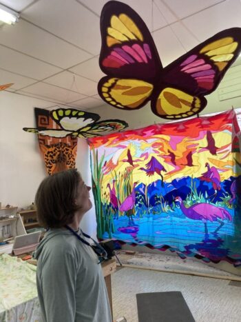 Person looking up at large, colorful butterfly kite with a vibrant flamingo tapestry and art supplies in the background.