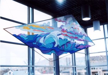 Colorful geometric glass artwork with ocean and whale design hangs in front of large windows in a modern building.
