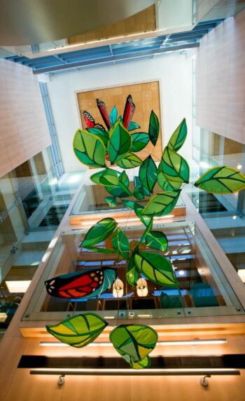 Large green leaf and butterfly sculptures hang in a glass atrium with natural light and wood-paneled walls.