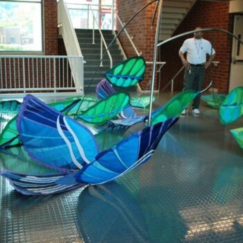 Colorful blue and green abstract sculptures are displayed indoors, with a man standing nearby and stairs in the background.