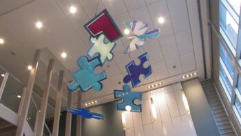 Large colored puzzle piece sculptures hang from the ceiling in a modern building with high windows and white walls.