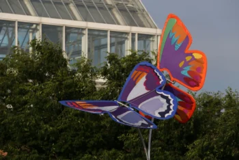 Two large, colorful butterfly sculptures are displayed outdoors near green trees and a glass greenhouse building.