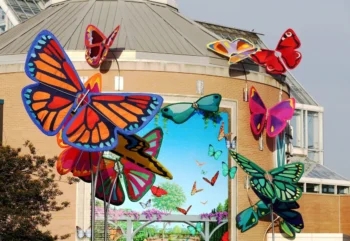 Oversized butterfly sculptures stand outside a round building with a butterfly and garden mural on its wall.