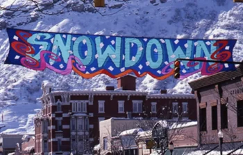 A colorful "Snowdown" banner hangs over a snowy street with historic brick buildings and mountains in the background.