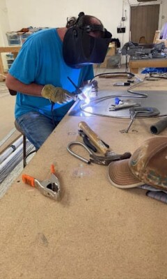 Person wearing a welding mask and gloves uses a torch to weld metal at a workbench with tools and a cap.