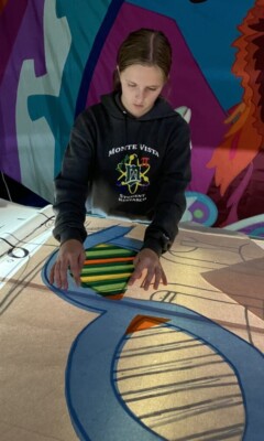 A person arranges colored tissue paper on a large, outlined artwork under strong lighting at a workspace.