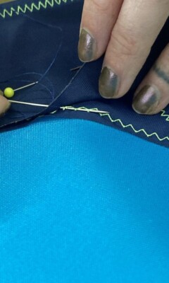 Close-up of hands with painted nails pinning blue fabric pieces together, showing visible stitching and zigzag seams.
