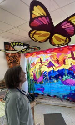 Person looking up at large butterfly decorations in a colorful art studio with a vibrant bird tapestry on the wall.